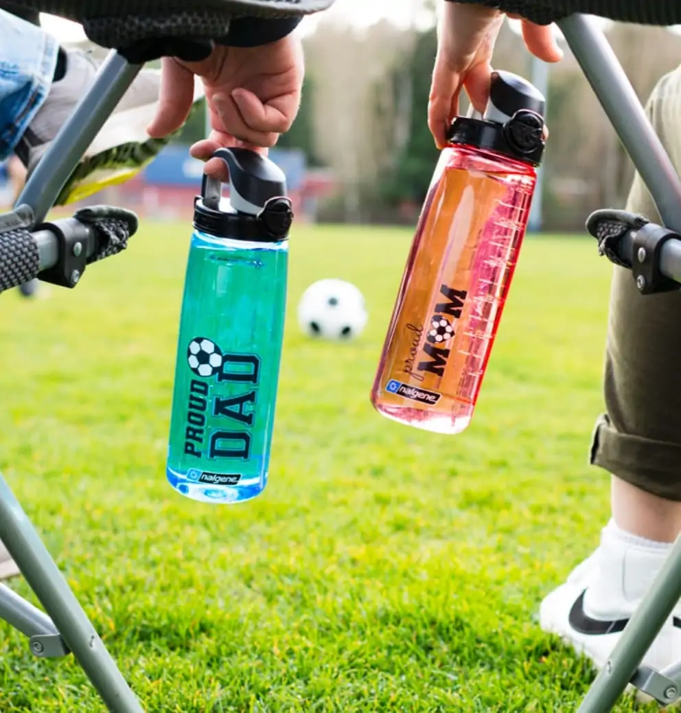 image of two personalized nalgene water bottles with "Proud Dad" and "Proud Mom". Looks to be two people holding them while sitting on a soccer field