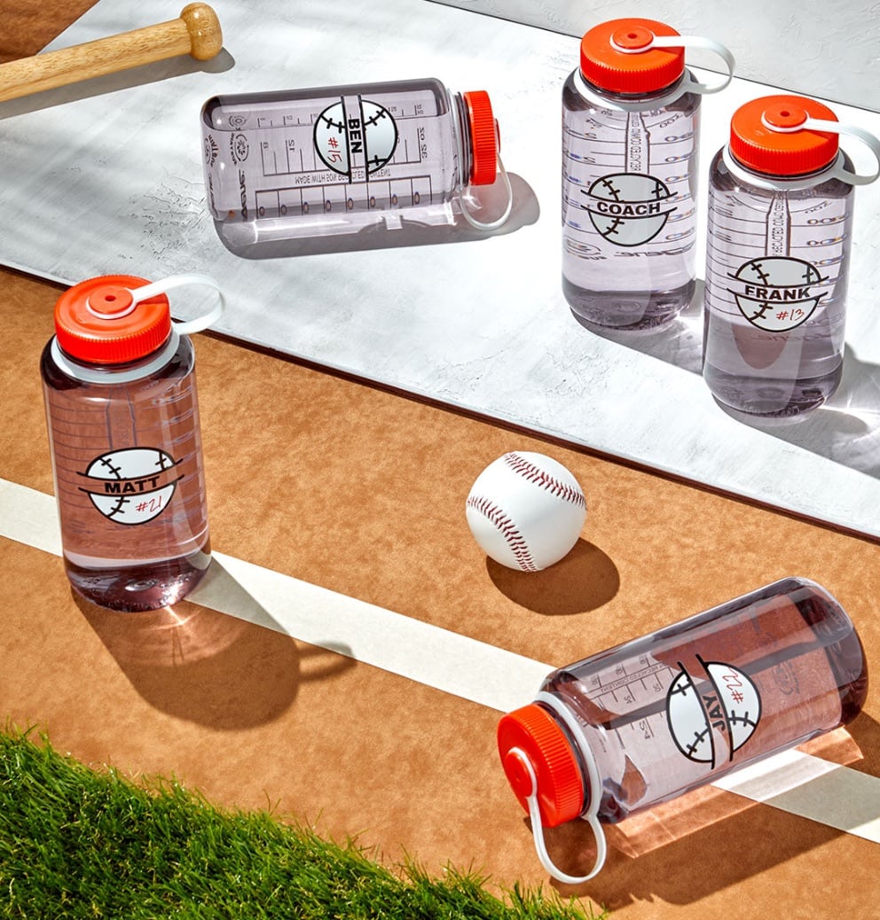 image of 5 nalgene water bottles with a baseball on the front and a person's name. They are all standing/laying on what looks to be a baseball field with a baseball in the shot and the handle of a baseball bat