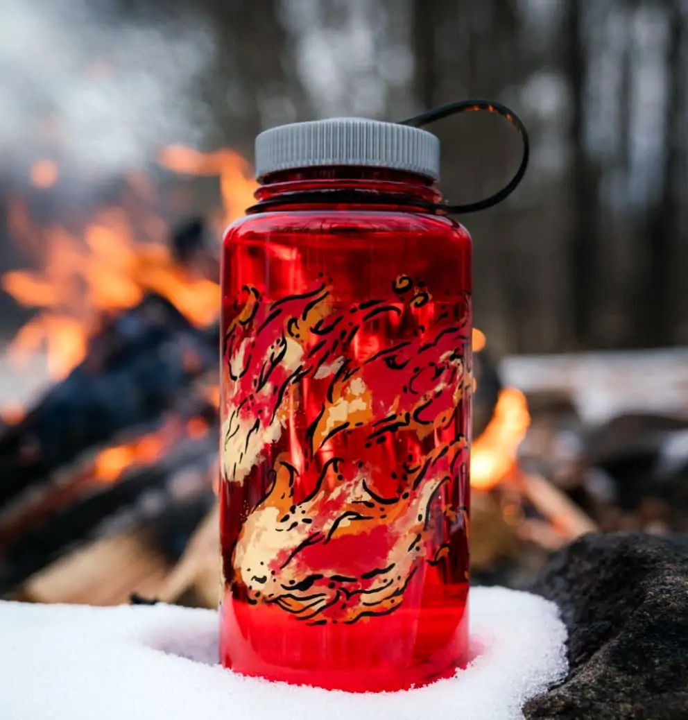 image of a nalgene water bottle sitting in some snow with a campfire in the background